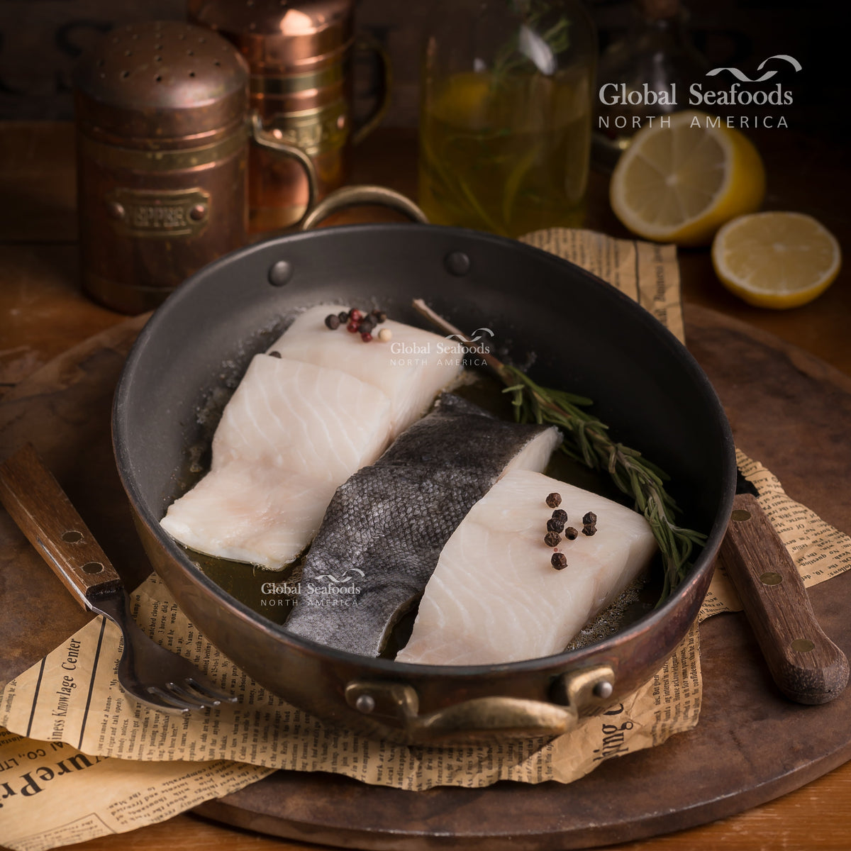 Two raw skin-on fish portions in a skillet with herbs and lemon nearby.