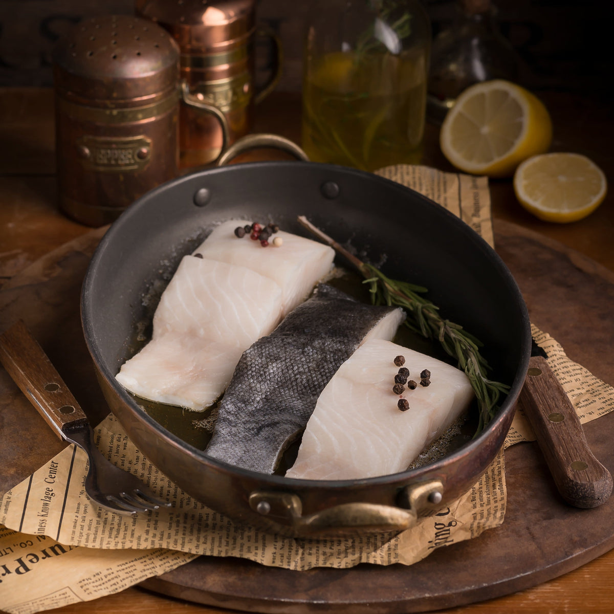 Two raw skin-on fish portions in a skillet with herbs and lemon nearby.