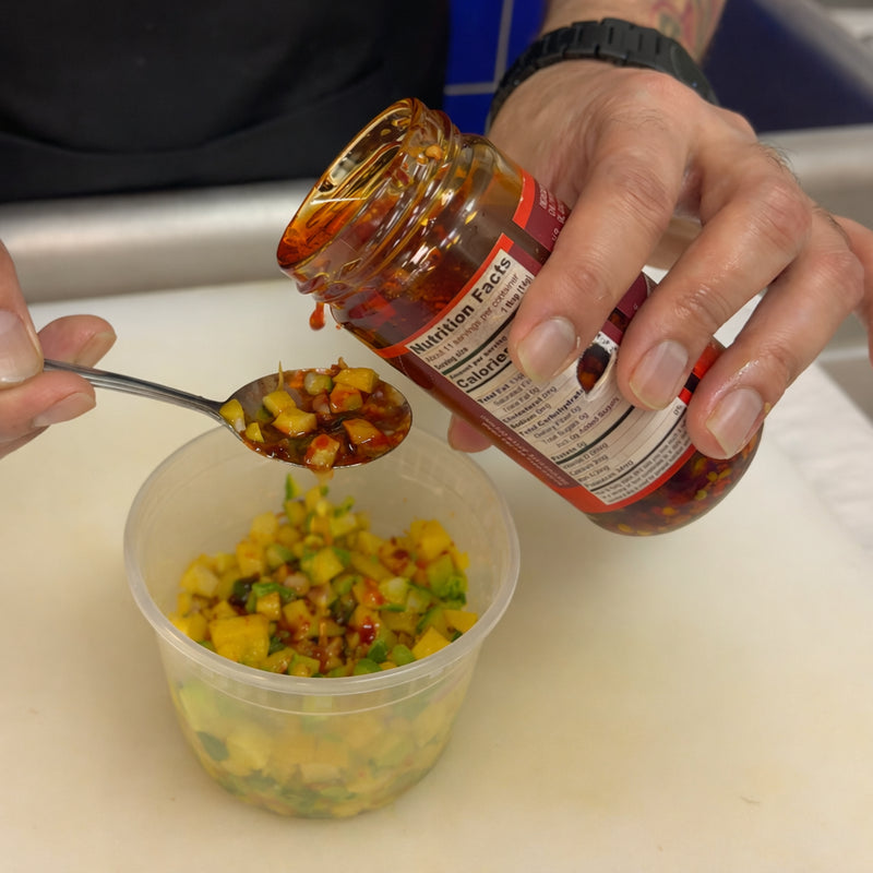 Adding a spoonful of chili condiment from a jar to diced mango salsa in a mixing container.