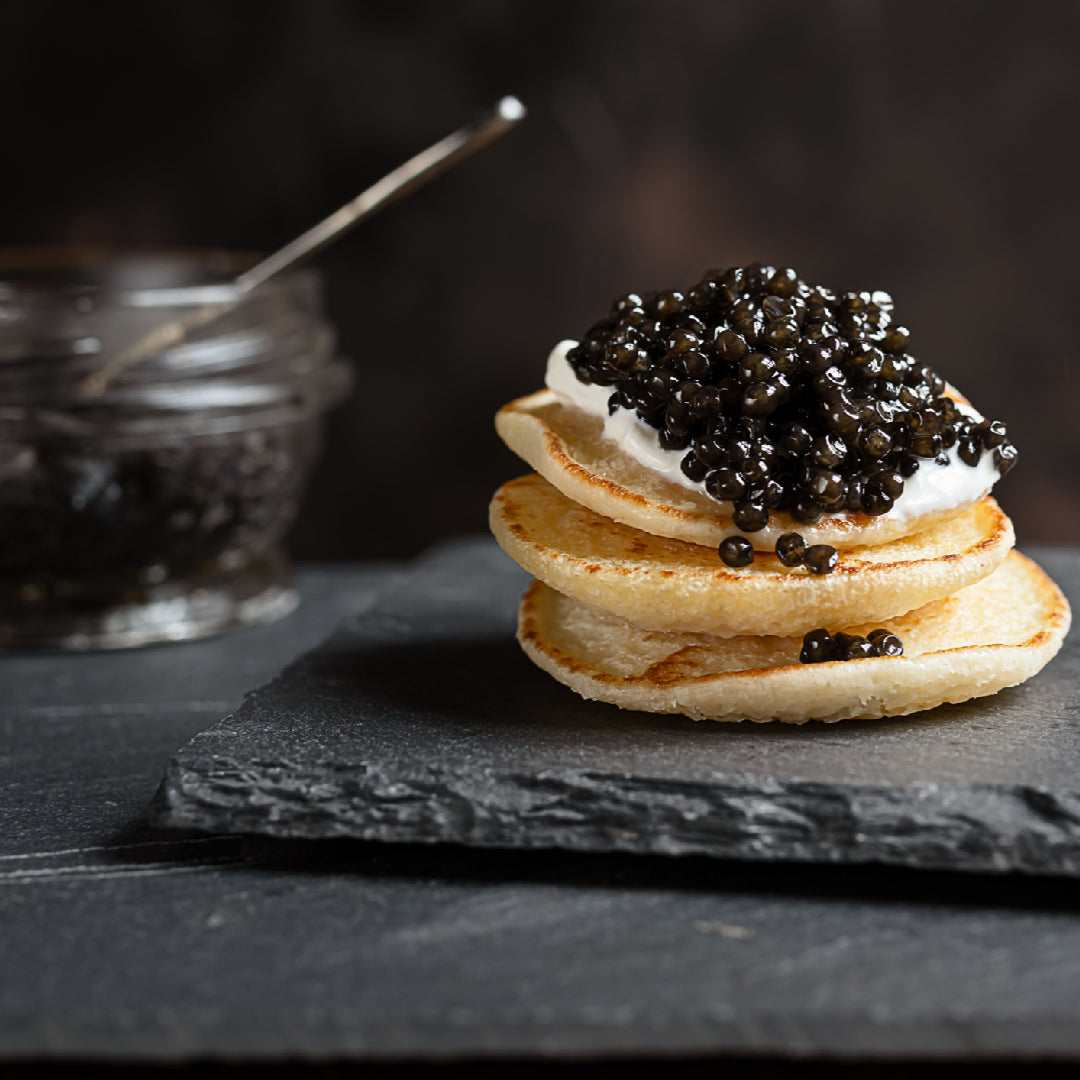 Open tin of premium Kaluga caviar with large glossy dark gray pearls displayed on crushed ice with a mother-of-pearl spoon and champagne flute