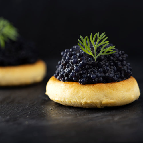 Small glossy black Bowfin caviar pearls served on blinis with crème fraîche and fresh herbs on a dark slate board