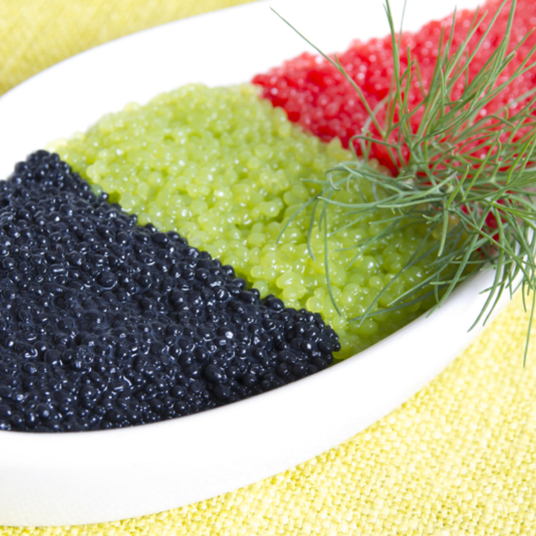Small jar of grayish-black paddlefish caviar pearls served on a white plate with blinis and crème fraîche alongside a mother-of-pearl spoon
