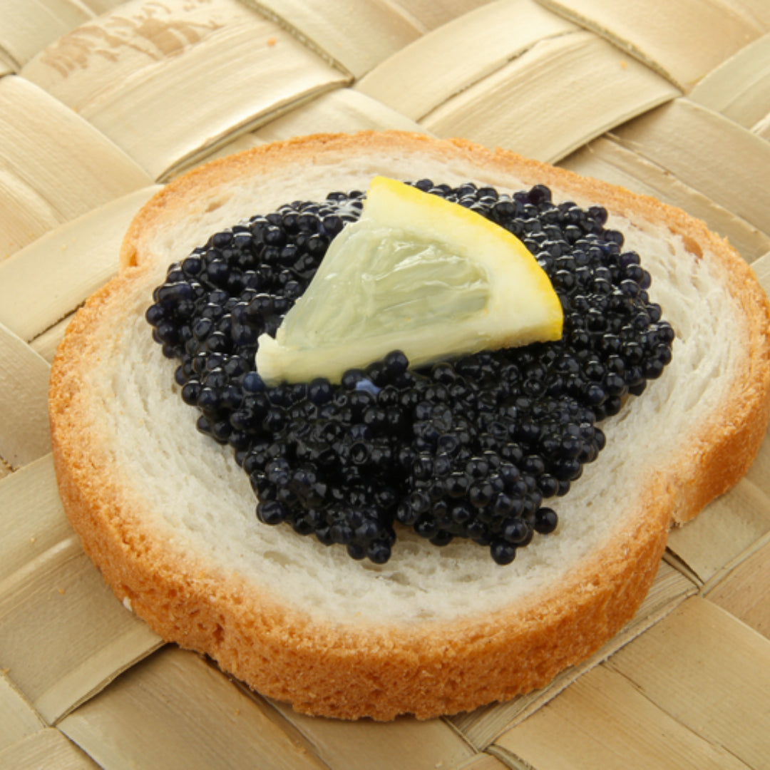 Large glistening Kaluga caviar pearls in a mother-of-pearl spoon resting on crushed ice beside blinis and crème fraîche on a dark slate board