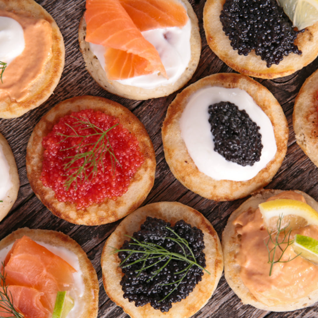 Mini blinis topped with crème fraîche and glistening black Kaluga caviar pearls arranged on a slate board with a mother-of-pearl spoon