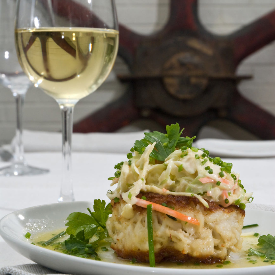A plate of golden-crusted crab cakes alongside a chilled glass of Sauvignon Blanc — one of the classic wine pairings for crab, where the wine's acidity cuts through the richness of the crab cake and amplifies the sweetness of the crab meat
