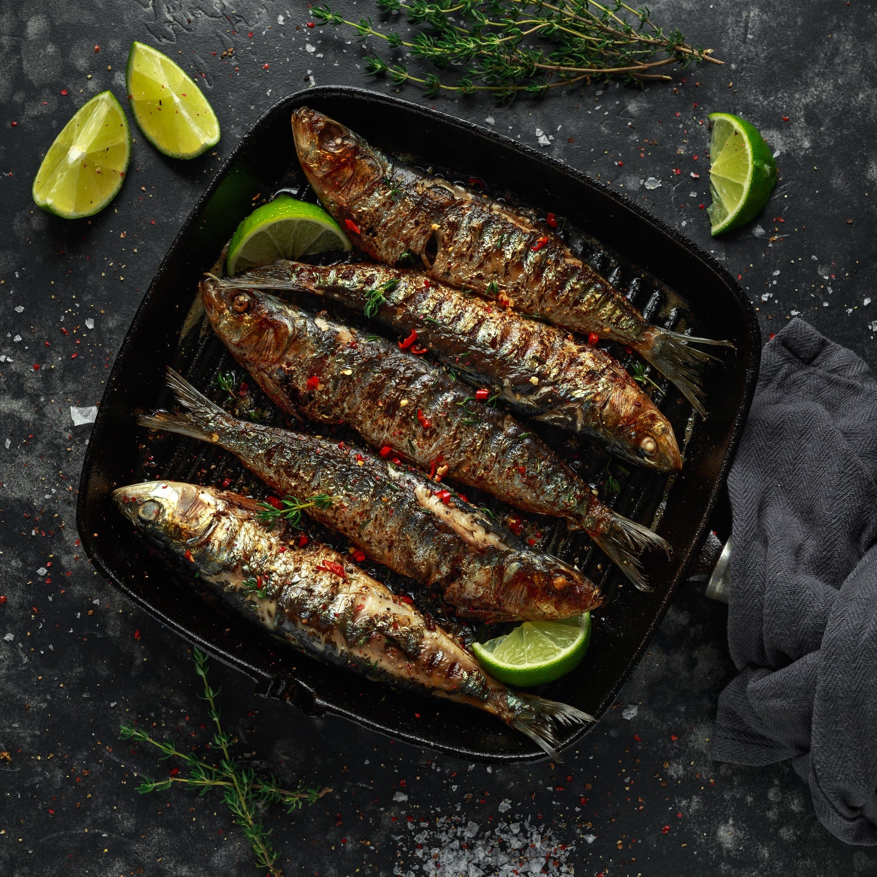 Grilled fresh sardines on a plate with lemon wedges, herbs, and olive oil on a rustic wooden table