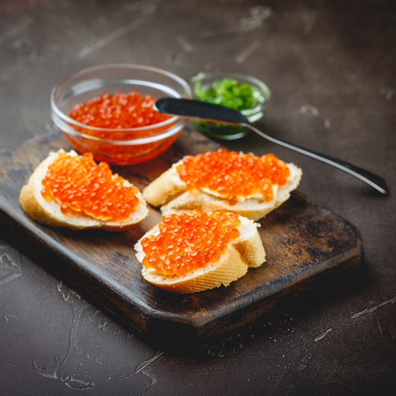 Side-by-side comparison of vibrant orange ikura salmon roe and dark sturgeon caviar in glass bowls on a marble surface