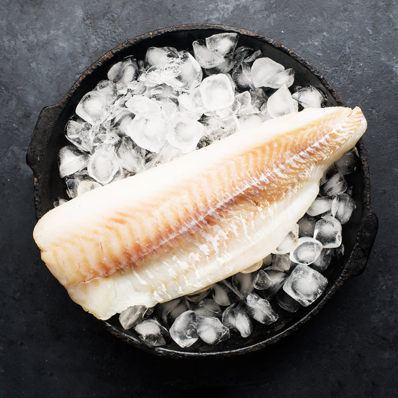 Fresh lingcod fillet with blue-green flesh on a cutting board with herbs and lemon
