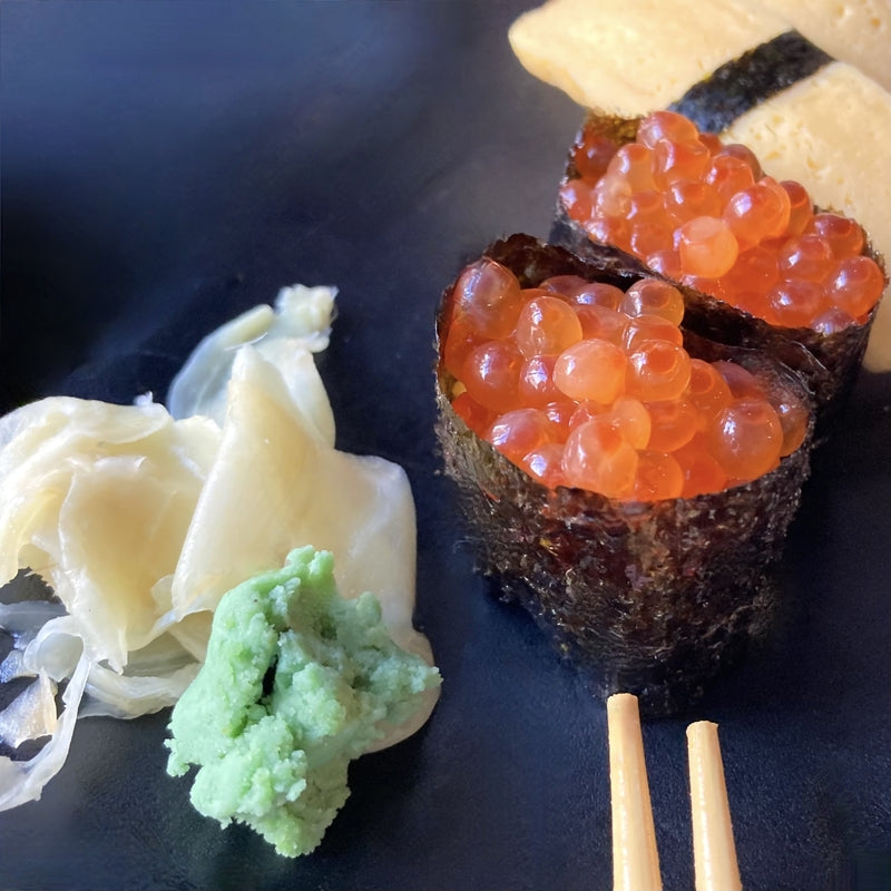 Elegantly plated ikura nigiri sushi arranged in a circular pattern on a dark slate board, garnished with shiso leaves and edible flowers