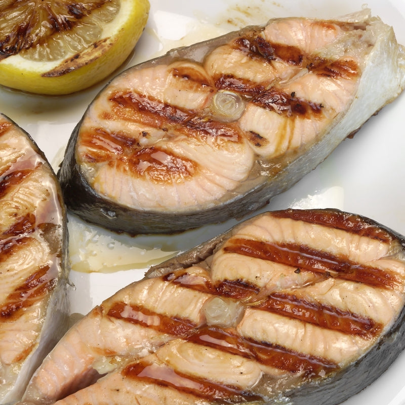Fresh fish fillets sizzling on a hot grill with char marks, surrounded by lemon slices and fresh herbs