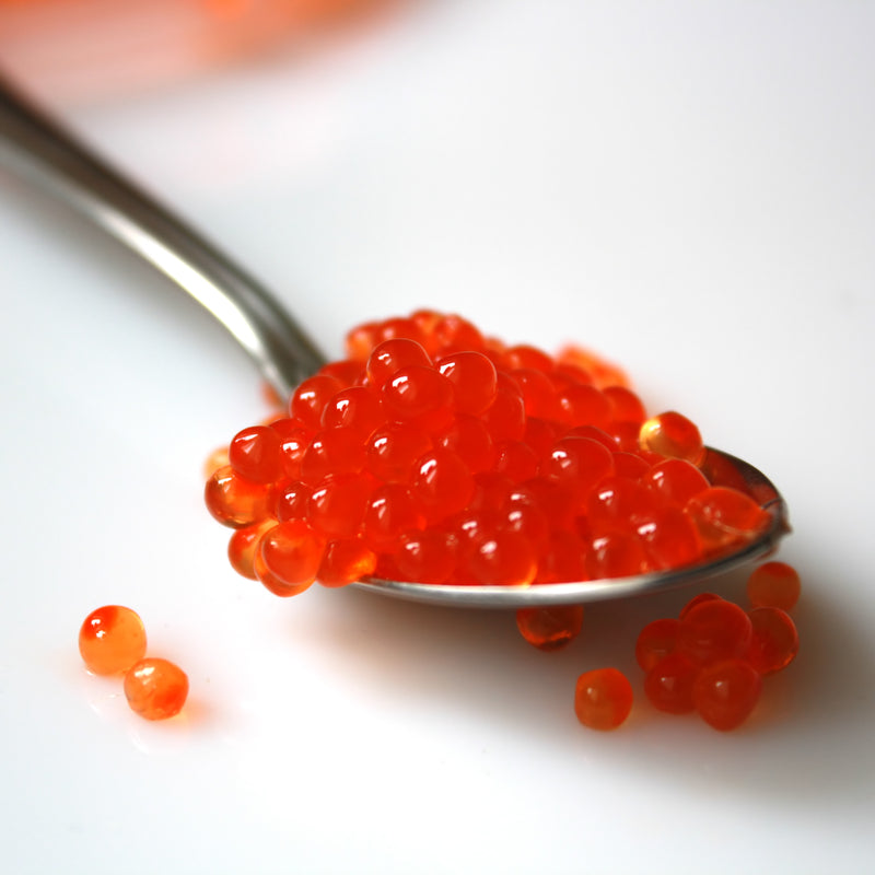Vibrant orange salmon roe pearls in a glass jar surrounded by fresh dill and lemon slices on a wooden surface