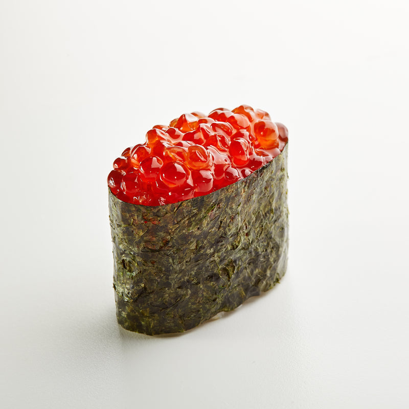 Bright orange ikura salmon roe in a small bowl beside a sushi roll and chopsticks on a wooden surface