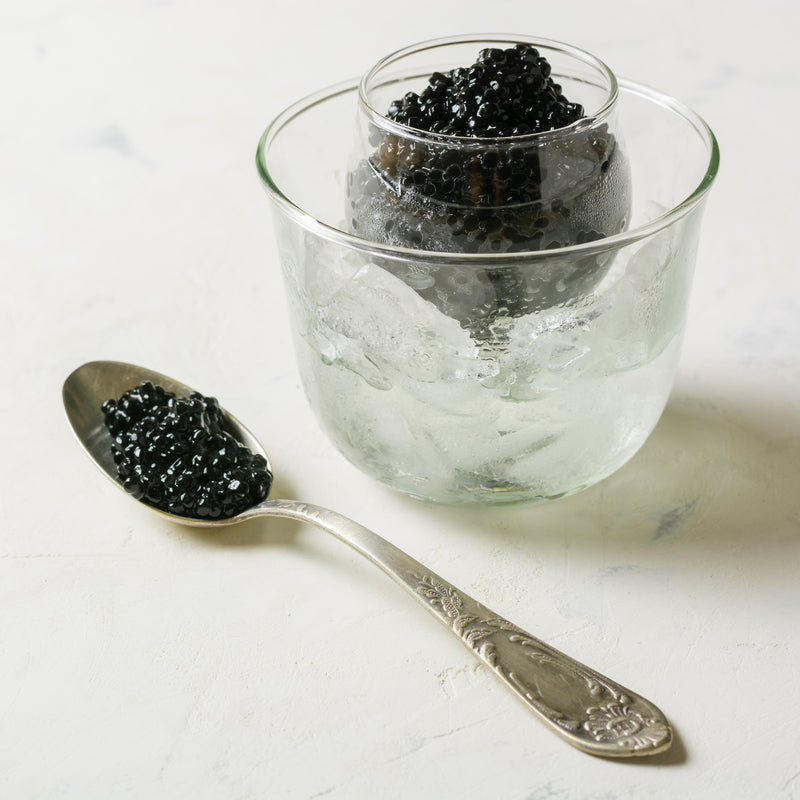 Romanoff caviar pearls in a tin on crushed ice with blinis, crème fraîche, and a mother-of-pearl spoon on a dark surface