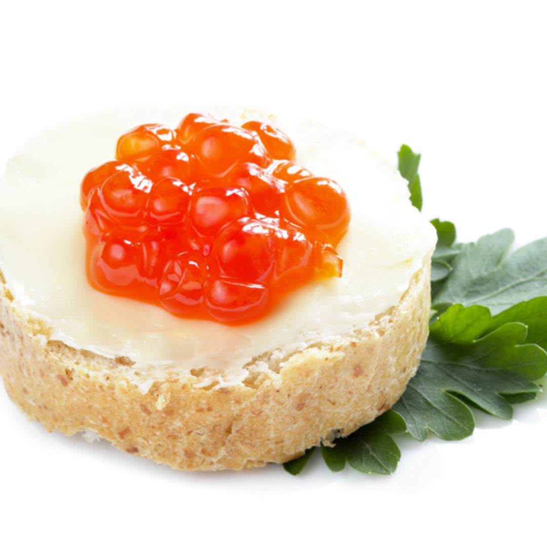 Glistening orange cured salmon roe pearls served in a small glass jar alongside toast points with cream cheese and fresh dill on a marble surface