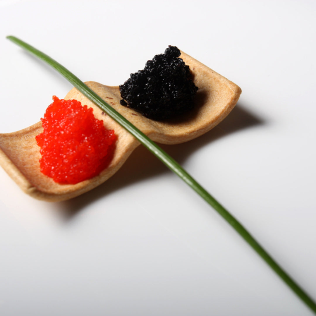 Side-by-side comparison of tobiko flying fish roe in orange and ikura salmon roe in red-orange on a white ceramic plate with sushi