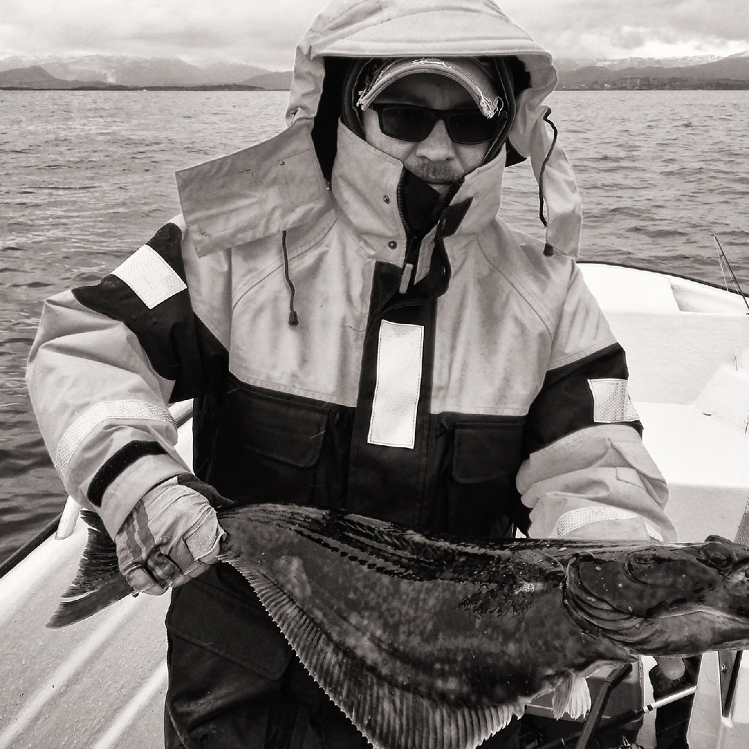 An angler on a fishing boat in a Norwegian fjord holding a large trophy halibut with dramatic mountain scenery in the background