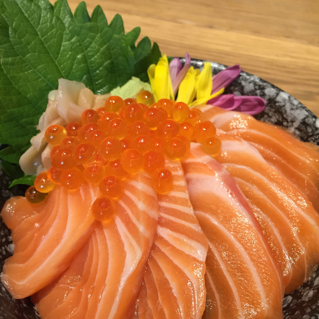 Ikura donburi in a ceramic bowl: seasoned sushi rice topped with vibrant orange salmon roe, shredded nori, sesame seeds, and shiso leaves