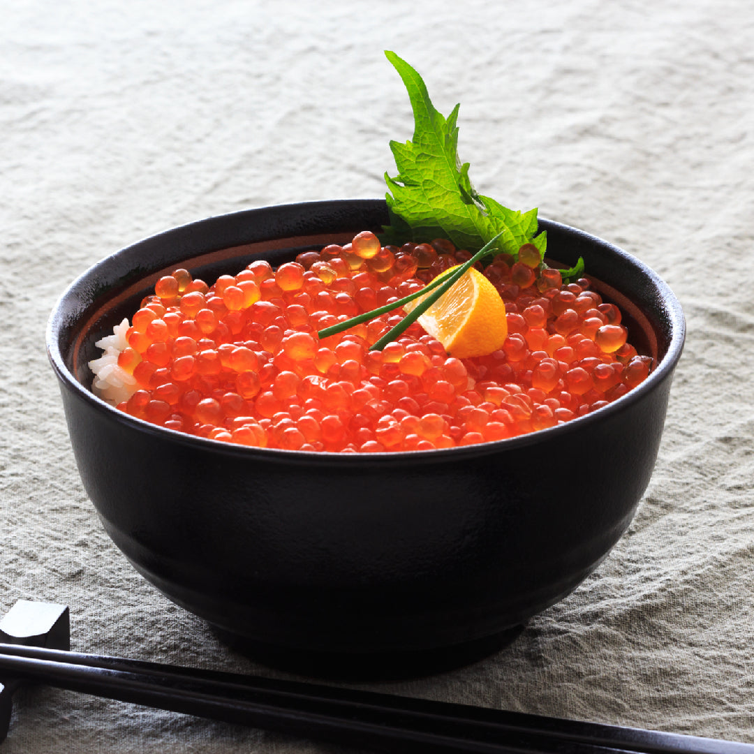 Triangular ikura onigiri rice balls wrapped in nori on a wooden board, filled with bright orange salmon roe, garnished with sesame seeds and green onions