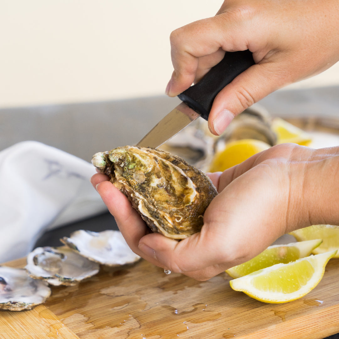 Oyster shucking knife inserted into the hinge of a fresh oyster on a wooden board