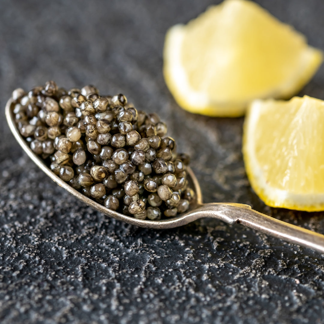 Osetra caviar tin resting on crushed ice next to a mother-of-pearl spoon, blini, and crème fraîche on a marble surface