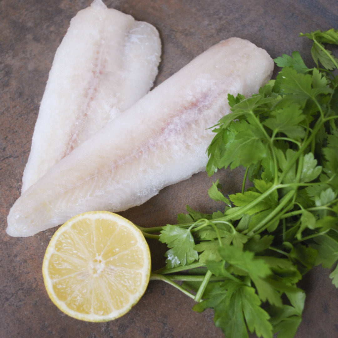 Grilled pollock fillet on a plate with lemon slices, fresh herbs, and roasted vegetables