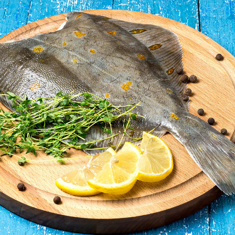 Whole flatfish with orange spots on a wooden board beside lemon slices and herbs