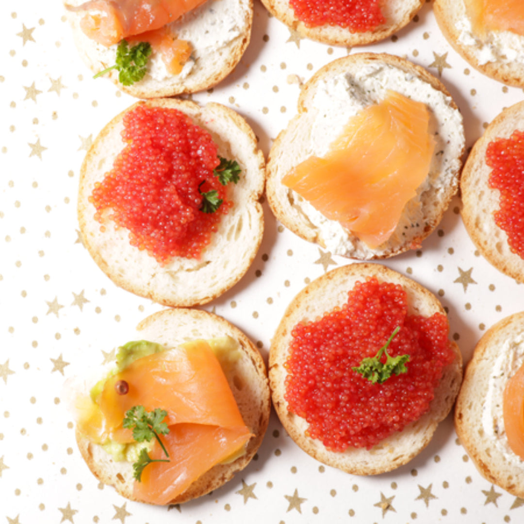 Vibrant orange-red salmon caviar pearls served in a small glass bowl with blinis and crème fraîche on a dark wooden board