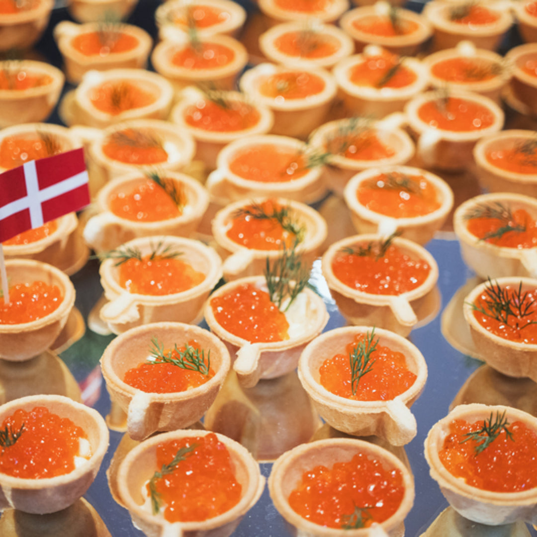 Vibrant orange salmon roe pearls served in a glass bowl on crushed ice alongside blinis, sour cream, and fresh dill on a white marble surface
