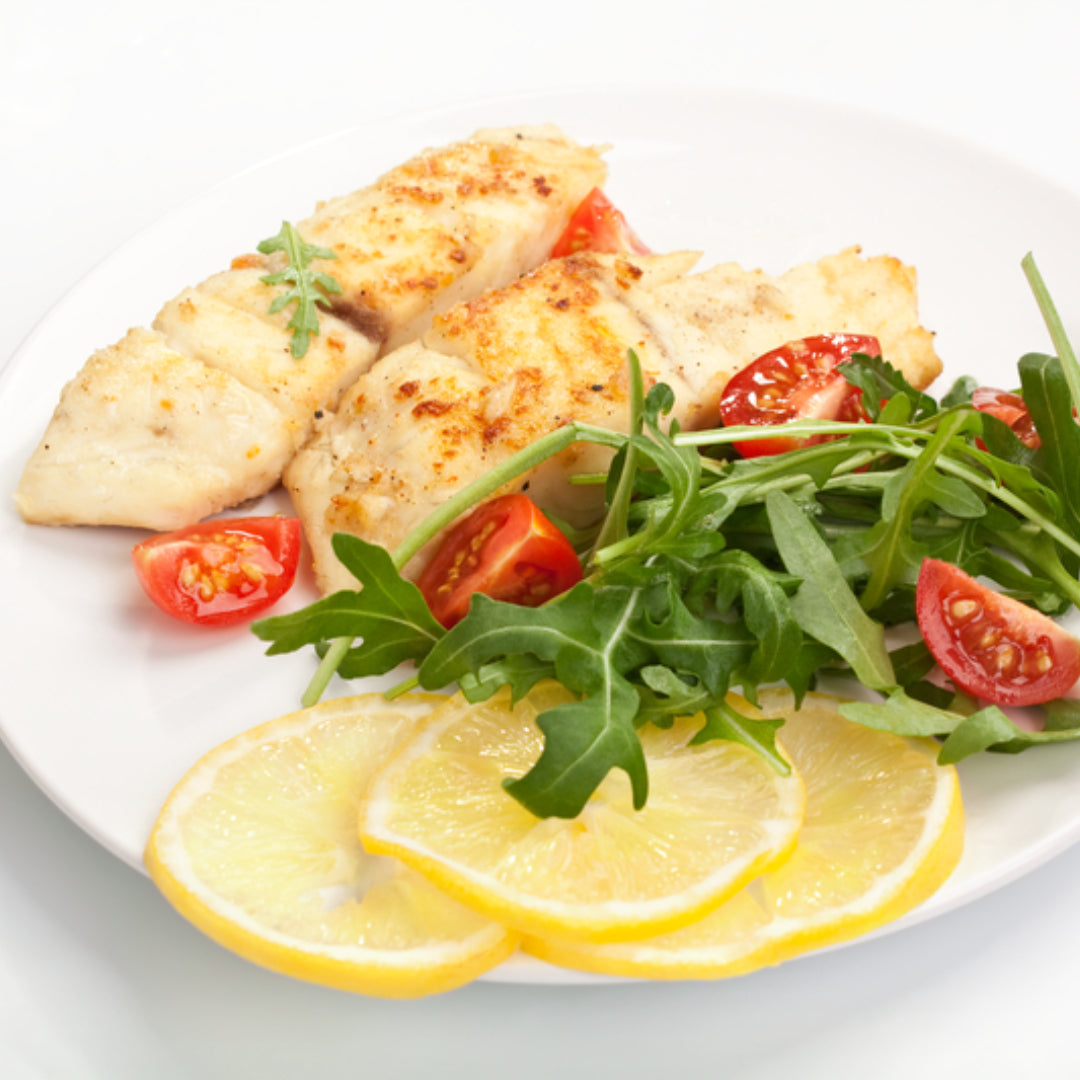 Cooked white fish with leafy greens, cherry tomatoes and lemon slices on a plate.