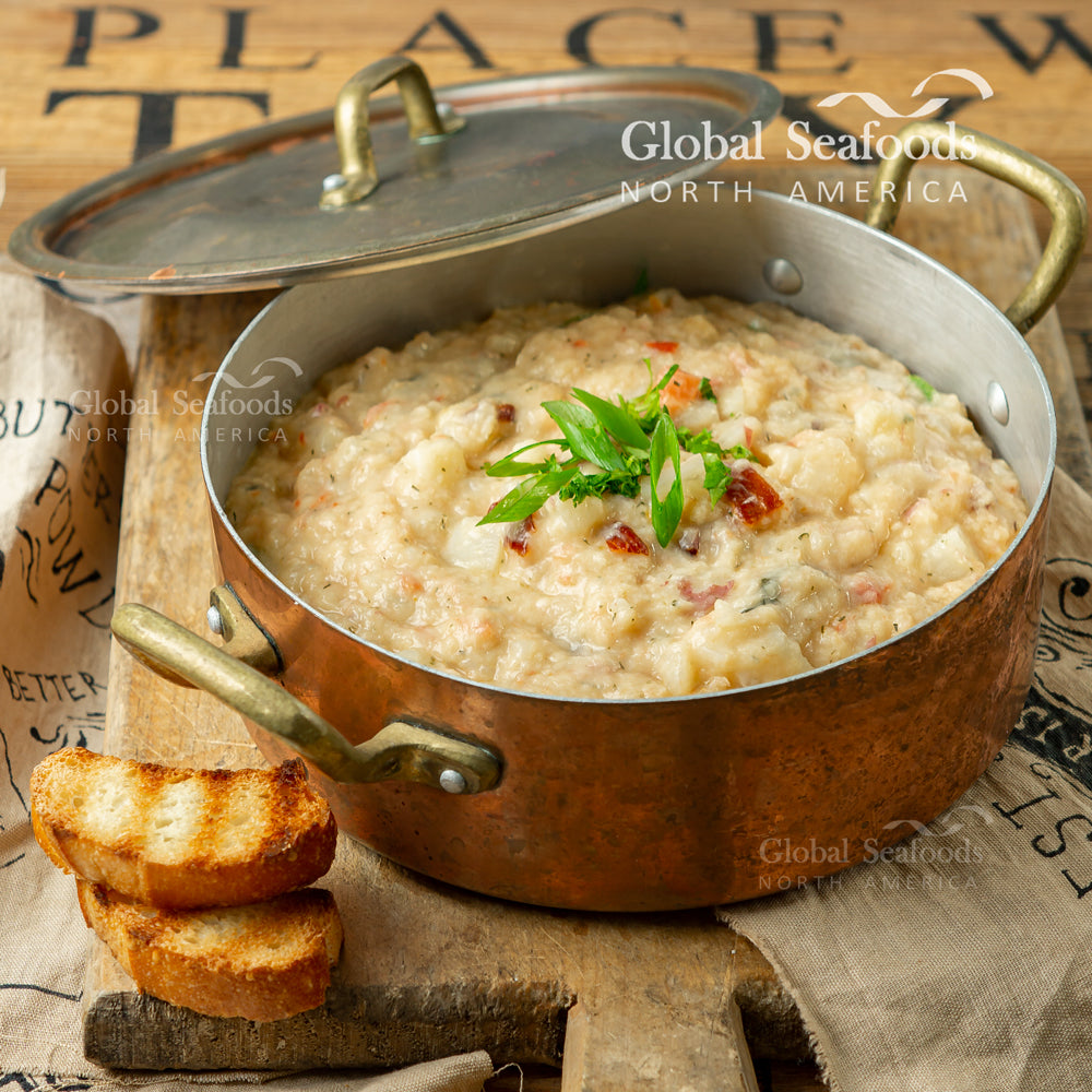 Salmon chowder in an outdoor copper metal pot with lid beside it, placed on a wooden cutting board with two toasted bread slices on the side