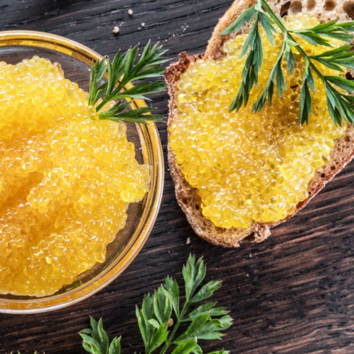 Golden Canadian pike roe in a glass bowl and on a slice of bread, garnished with herbs on a dark wooden surface