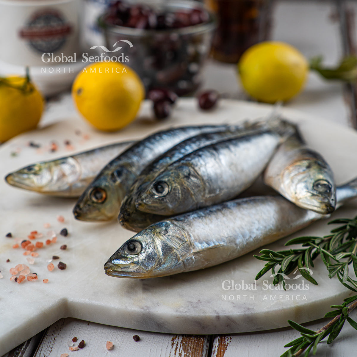 Whole sardines on a stone board beside rosemary and lemons