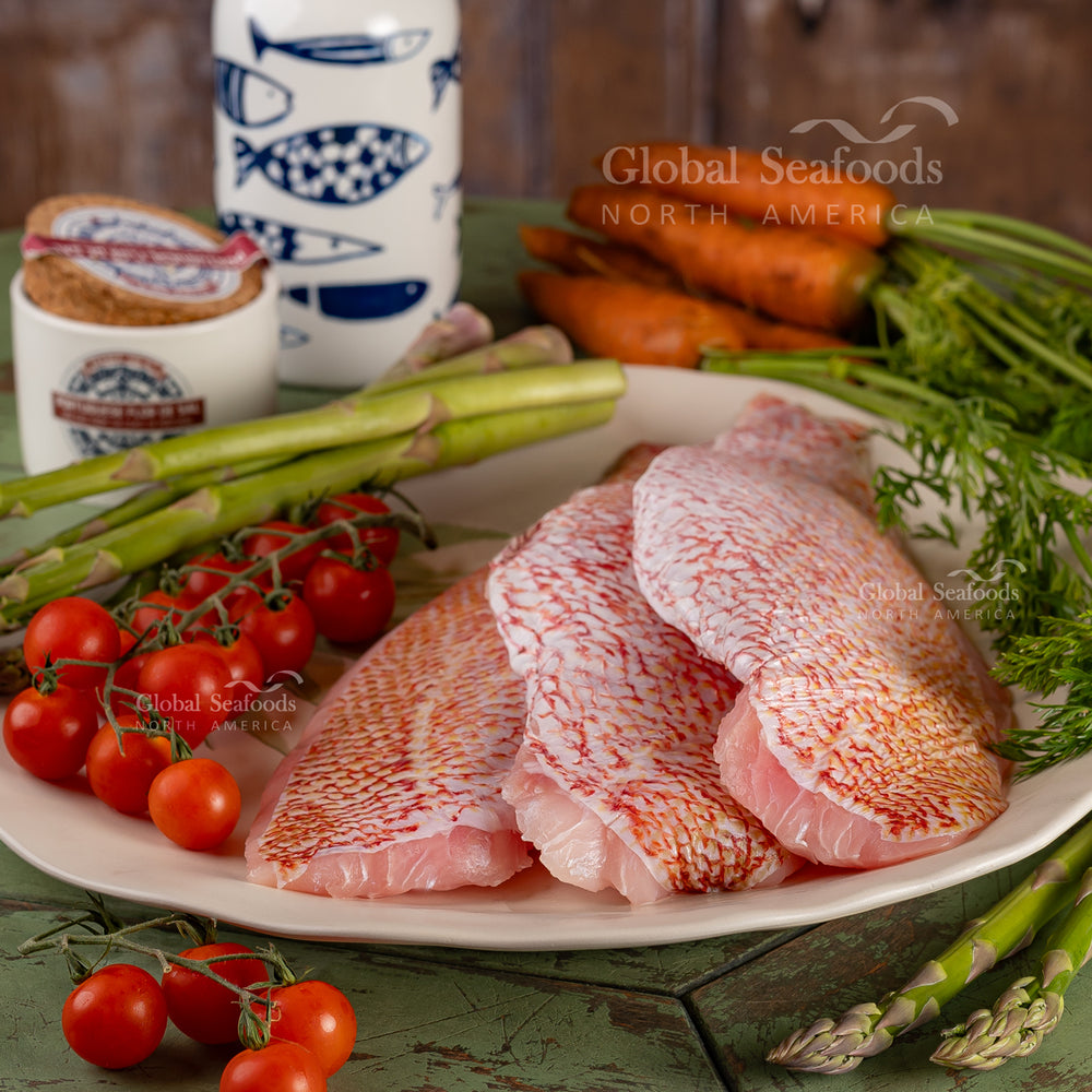 Fresh red snapper fillets with red skin served on a white plate, garnished with cherry tomatoes, asparagus, and carrots in the background – perfect seafood plating for grilling or gourmet dining