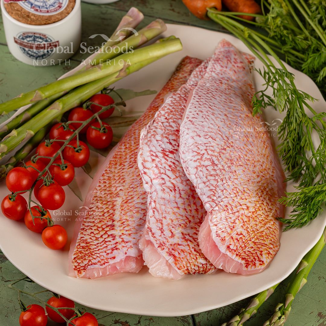 Close-up of three raw red snapper fillets, skin side up, on a white platter with asparagus and cherry tomatoes.