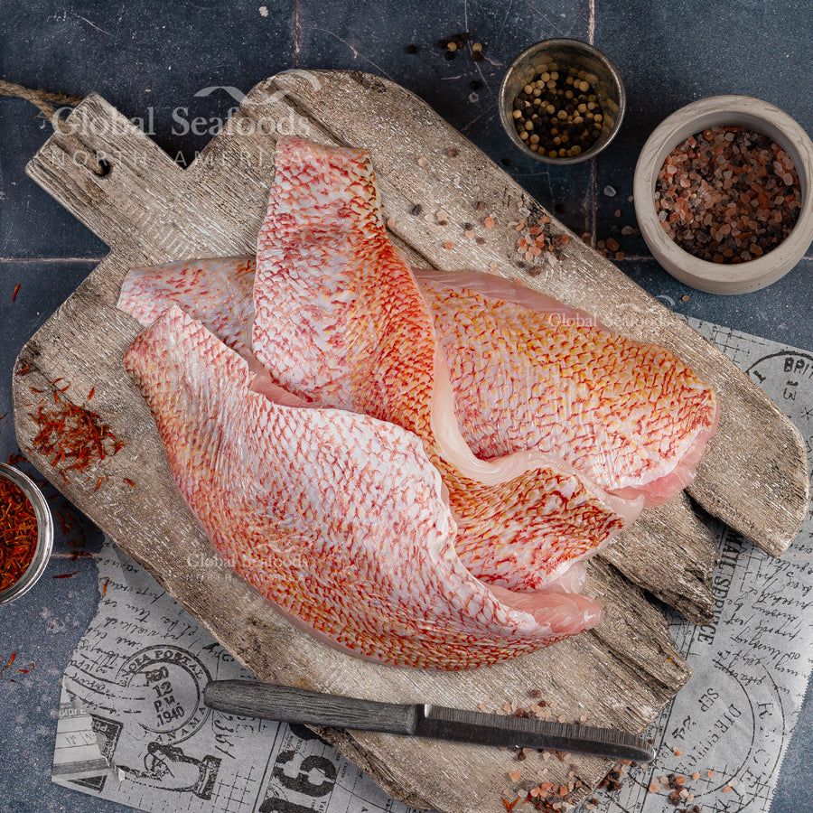 Raw skin-on red snapper fillets on a wooden cutting board beside coarse salt, peppercorns and a knife.