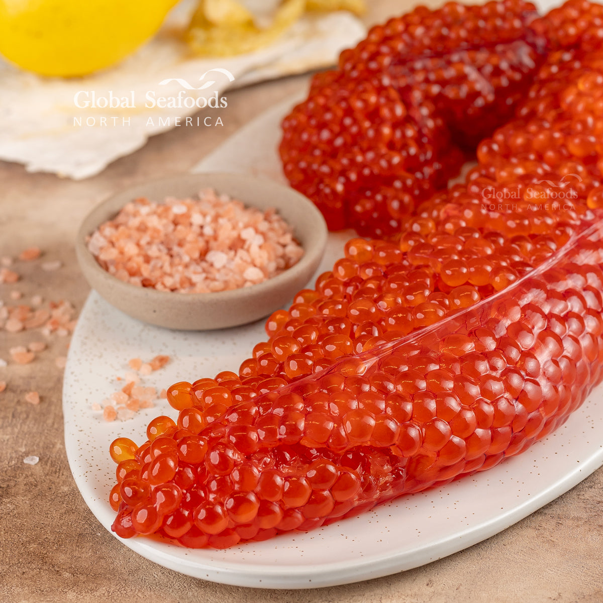 Close-up of unprocessed salmon roe skeins on a white plate beside a bowl of pink salt