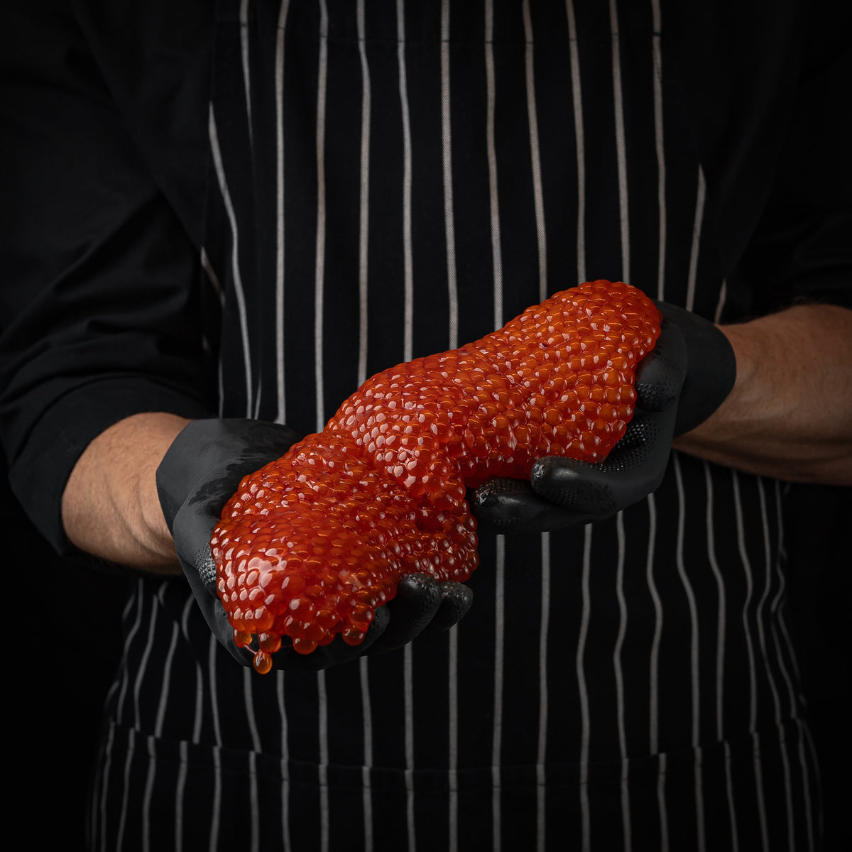 Person wearing black gloves holding an unprocessed salmon roe skein in front of a striped apron