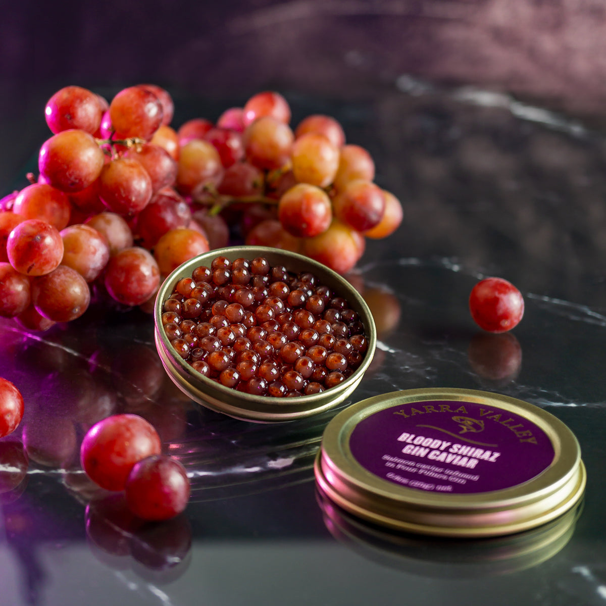 Bloody Shiraz gin-infused salmon roe in an open tin with red grapes and a labeled lid on a dark reflective surface