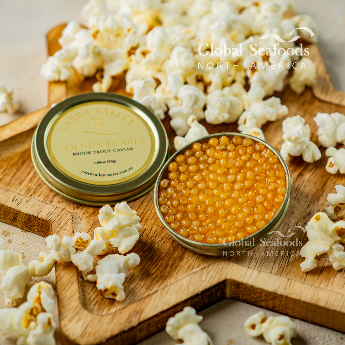 Open tin of golden brook trout caviar beside its labeled lid, surrounded by popcorn on a wooden cutting board