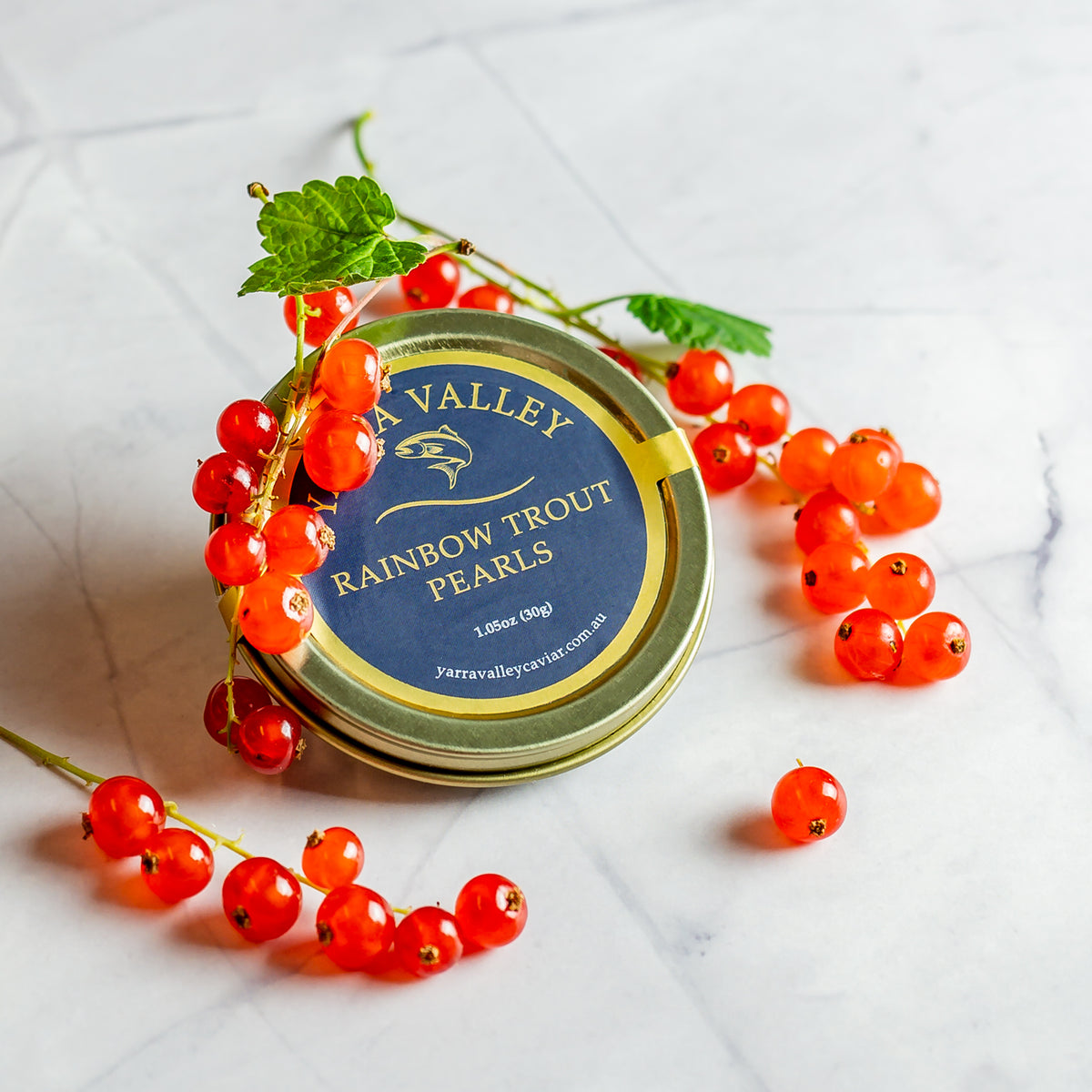Closed 30 g tin of Yarra Valley Rainbow Trout Pearls surrounded by red currants on a white marble surface