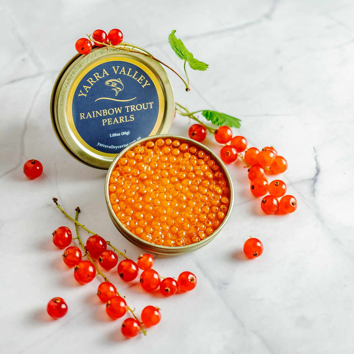 Open tin of Yarra Valley Rainbow Trout Pearls with its labeled lid and red currants on a white marble surface