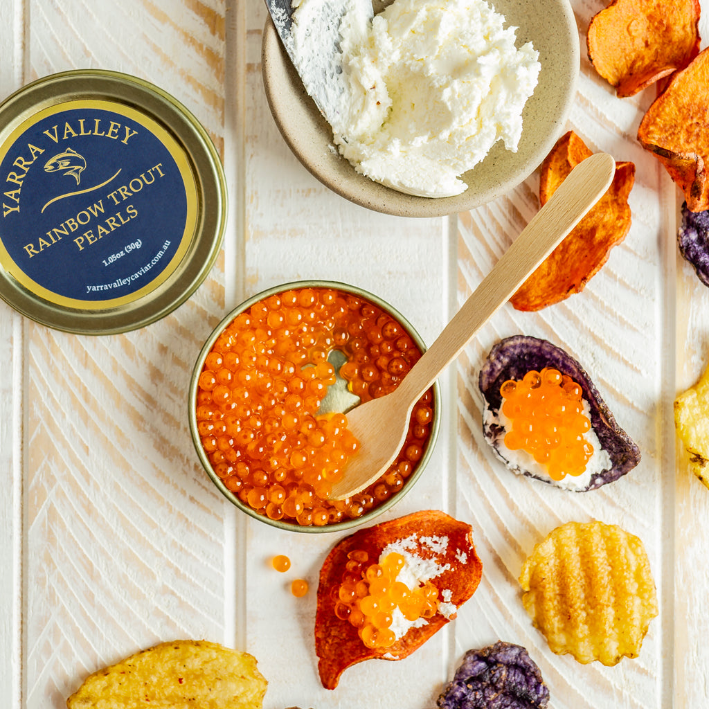 Rainbow trout roe in an open tin with a wooden spoon, beside vegetable chips topped with white cheese and roe