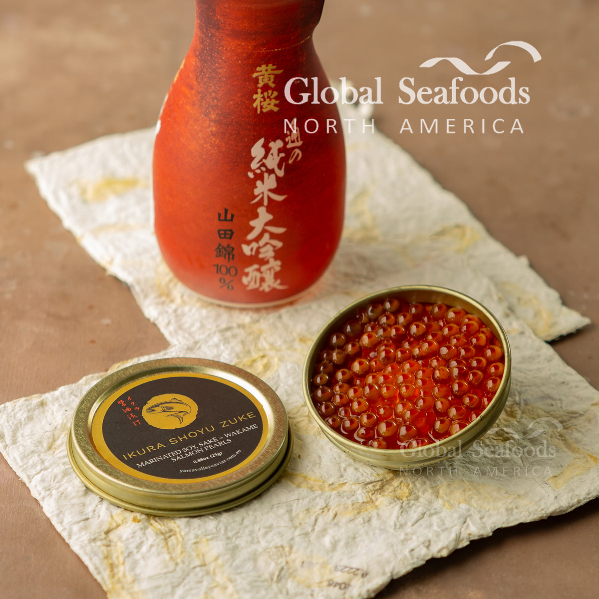 Open tin of Ikura Shoyu Zuke salmon roe and its labeled lid on textured paper beside a red sake bottle