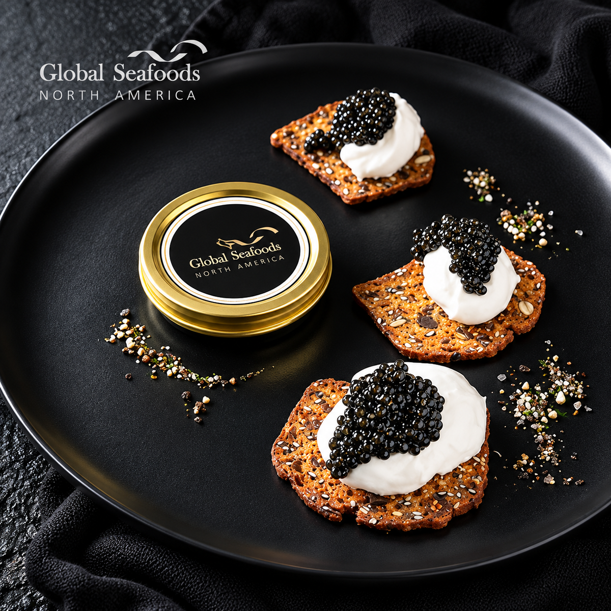 Three crackers topped with cream and Beluga hybrid caviar beside a closed tin on a black plate