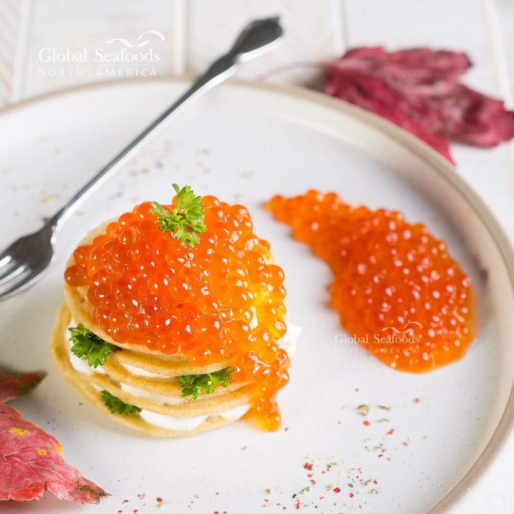 Bite-sized Russian-style blinis with sour cream filling and bright red Coho salmon roe (ikura) on top, plated on white ceramic
