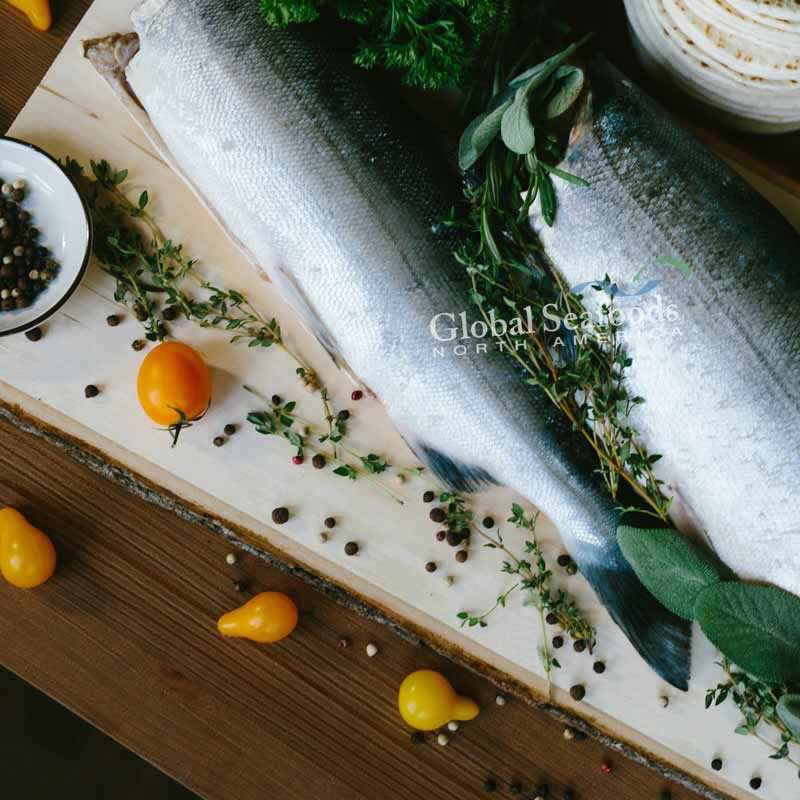 Two headless sockeye salmon on a wooden board with herbs and seasonings