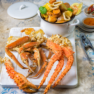 Spicy Cajun crab featuring snow crab and king crab clusters on a wooden board, with a pot in the background filled with boiled corn, sausages, squid rings, and shrimp for a hearty seafood boil presentation