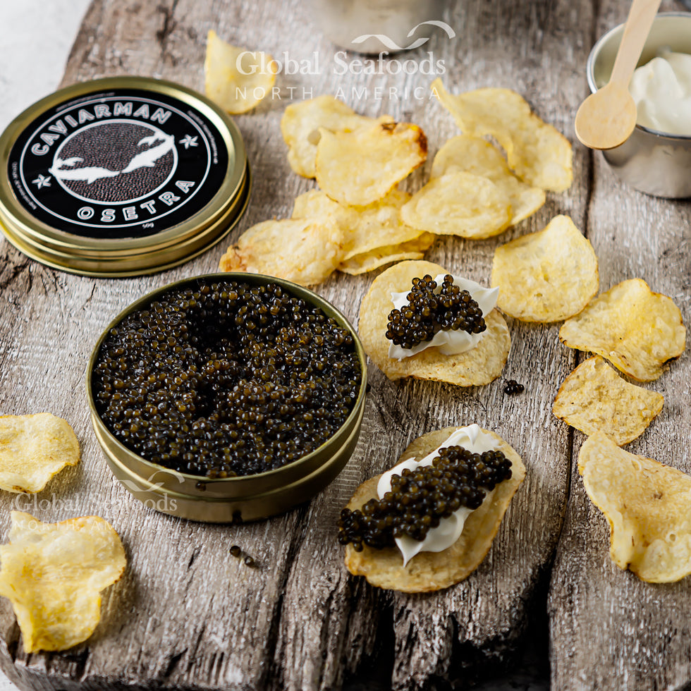 Russian Osetra caviar in an open tin beside potato chips topped with cream and caviar on a wooden surface
