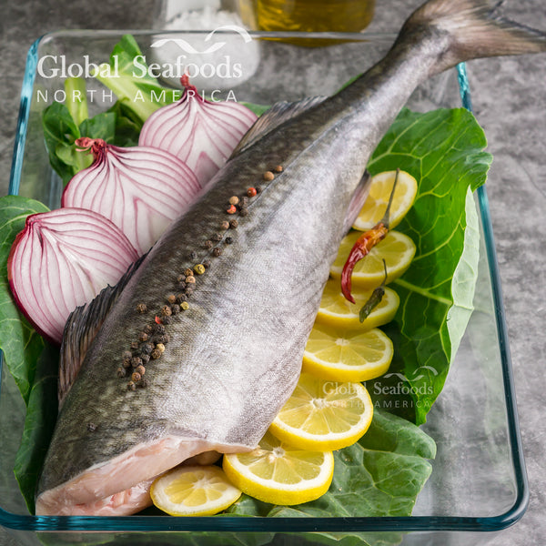 Headless sablefish in a glass tray with lemon, sliced red onion and greens.