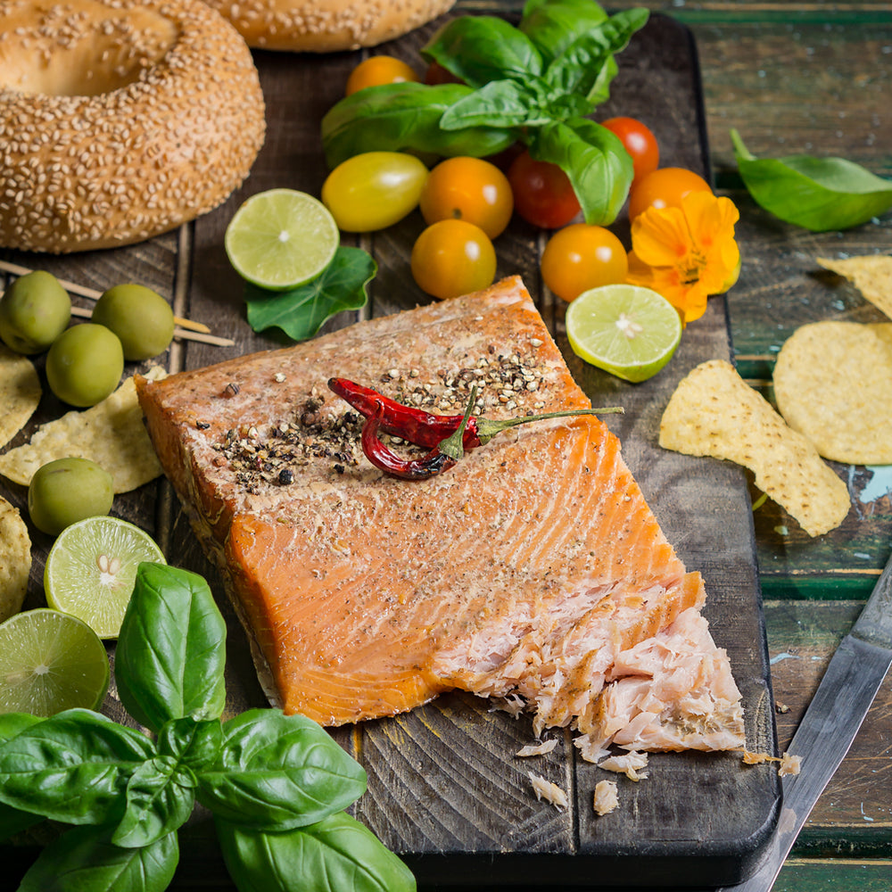Hot-smoked Alaskan king salmon fillet arranged on a wooden board with a fresh bagel, cherry tomatoes, lime slices, and olives for a vibrant deli-style serving